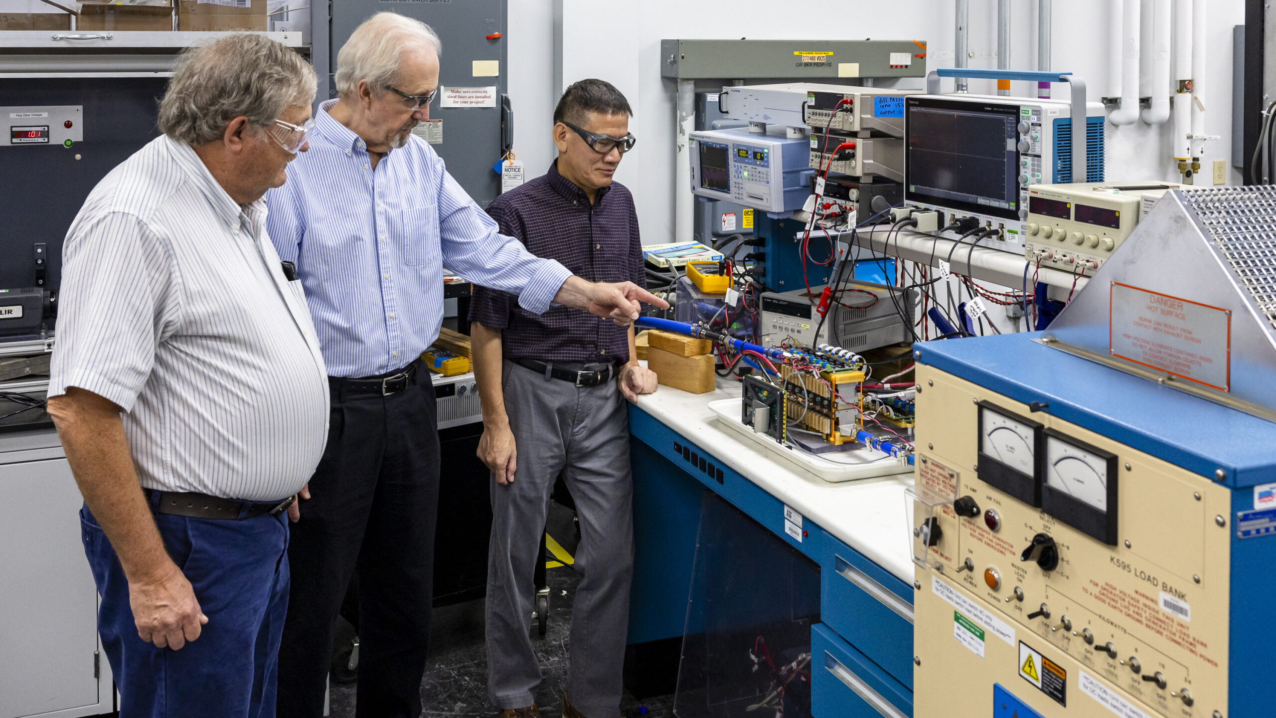 Three scientists wearing glasses are standing to the left, pointing at a bundle of electronics. There is a blue box with wooden cover to the right.