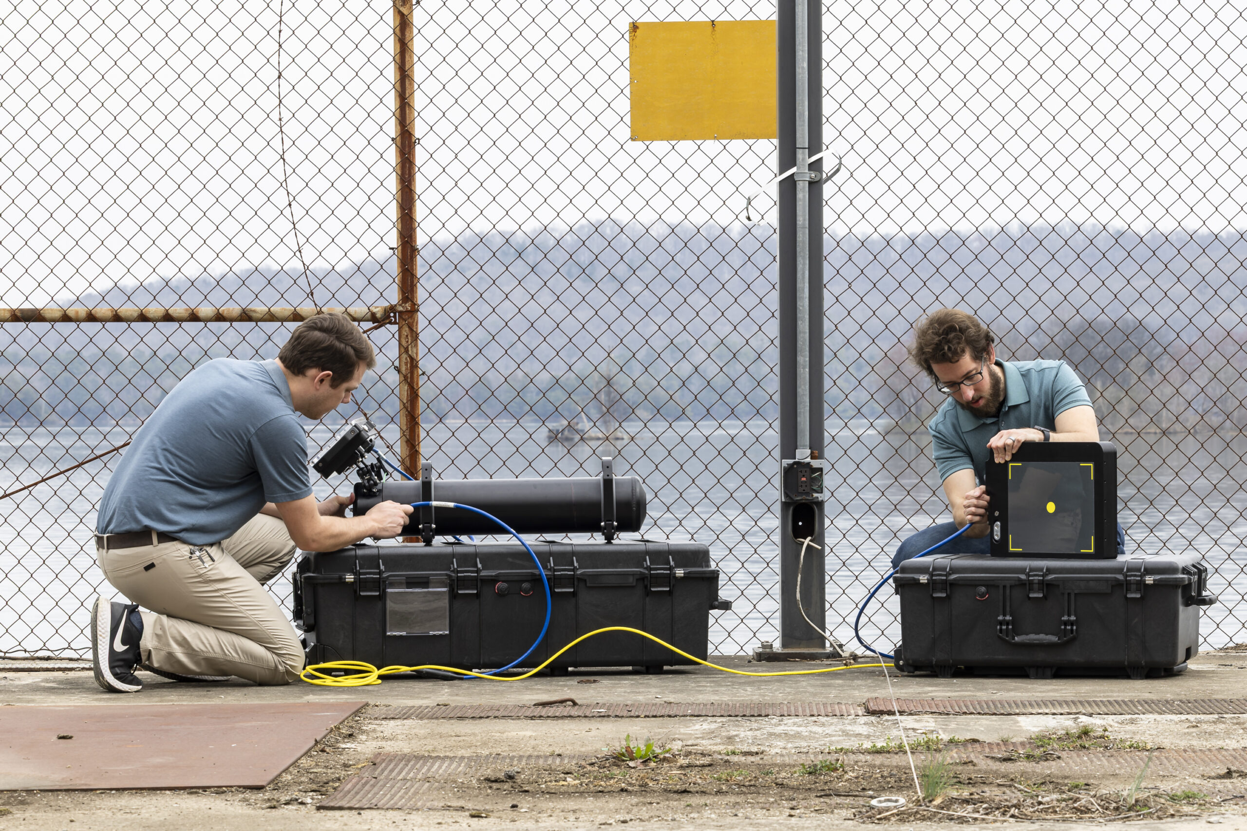Two researchers set up scientific equipment near a chain-link fence overlooking a body of water, connecting cables between a cylindrical instrument and a monitoring device mounted on portable cases.