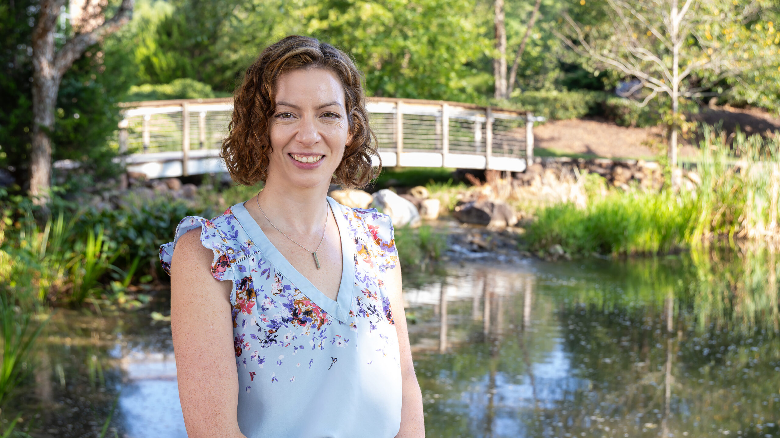 A woman with short curly hair smiles while standing outdoors in front of a pond. She is wearing a light blue floral sleeveless blouse and a necklace. Behind her, there is a small footbridge, green trees, and landscaped plants surrounding the water.