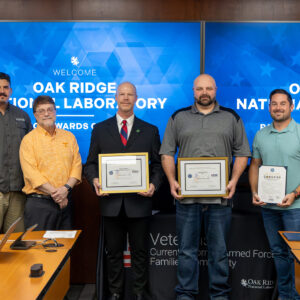 Seven men, including uniformed service members and ORNL staff, stand in front of Oak Ridge National Laboratory Patriot Awards Ceremony screens holding framed awards and a statement of support on February 27, 2026.