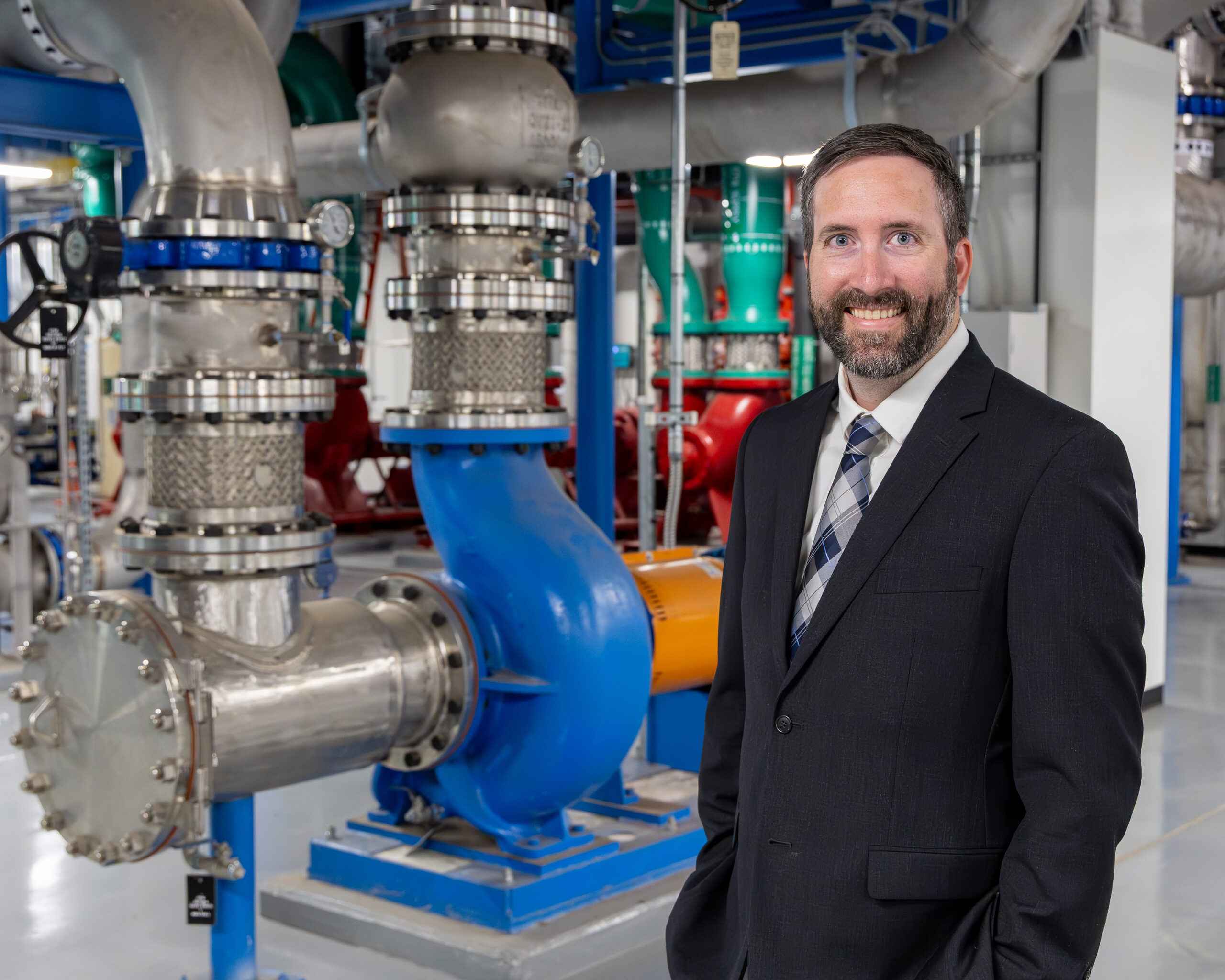 A man in a suit stands in front of large industrial piping and equipment inside a laboratory, smiling for a portrait.