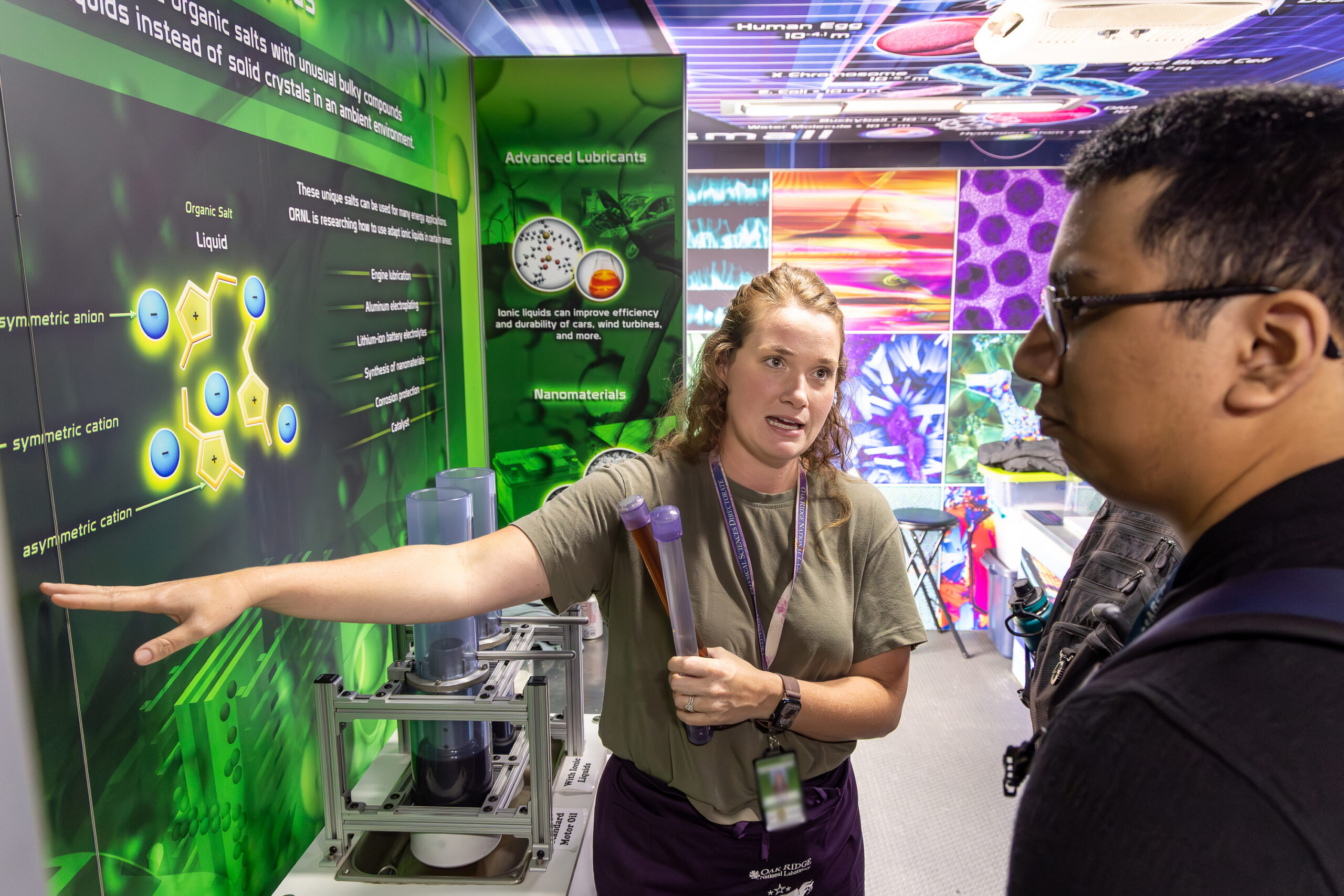 An Oak Ridge National Laboratory staff member explains a scientific exhibit about ionic liquids to a visitor inside a mobile research display. She points to a large illuminated diagram of molecular structures while holding demonstration tubes.