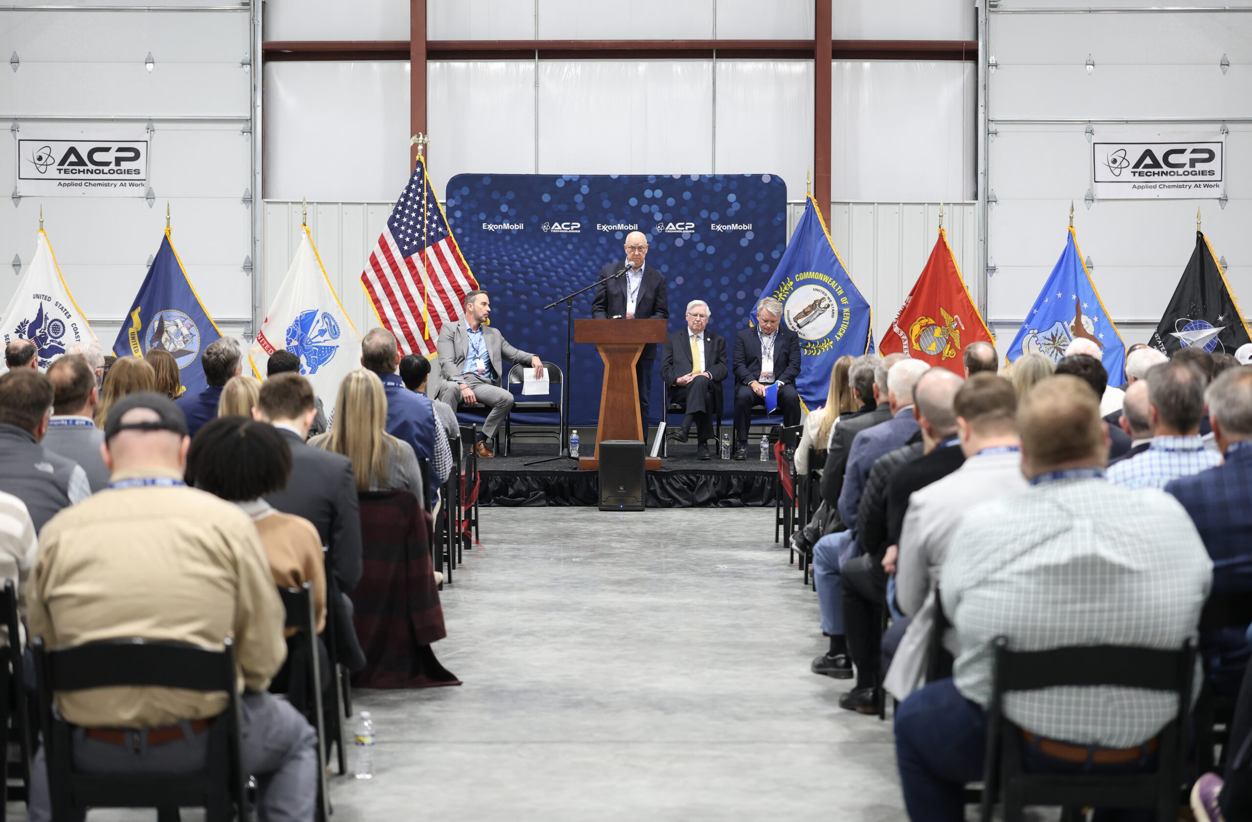 Audience seated in a large industrial facility listening to a speaker at a podium, with U.S. military and state flags displayed on stage behind him.