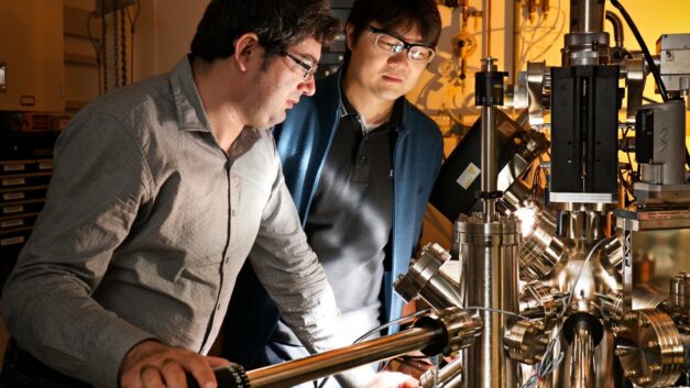 Two men in a room with an orange hue stare into a complex metallic machine