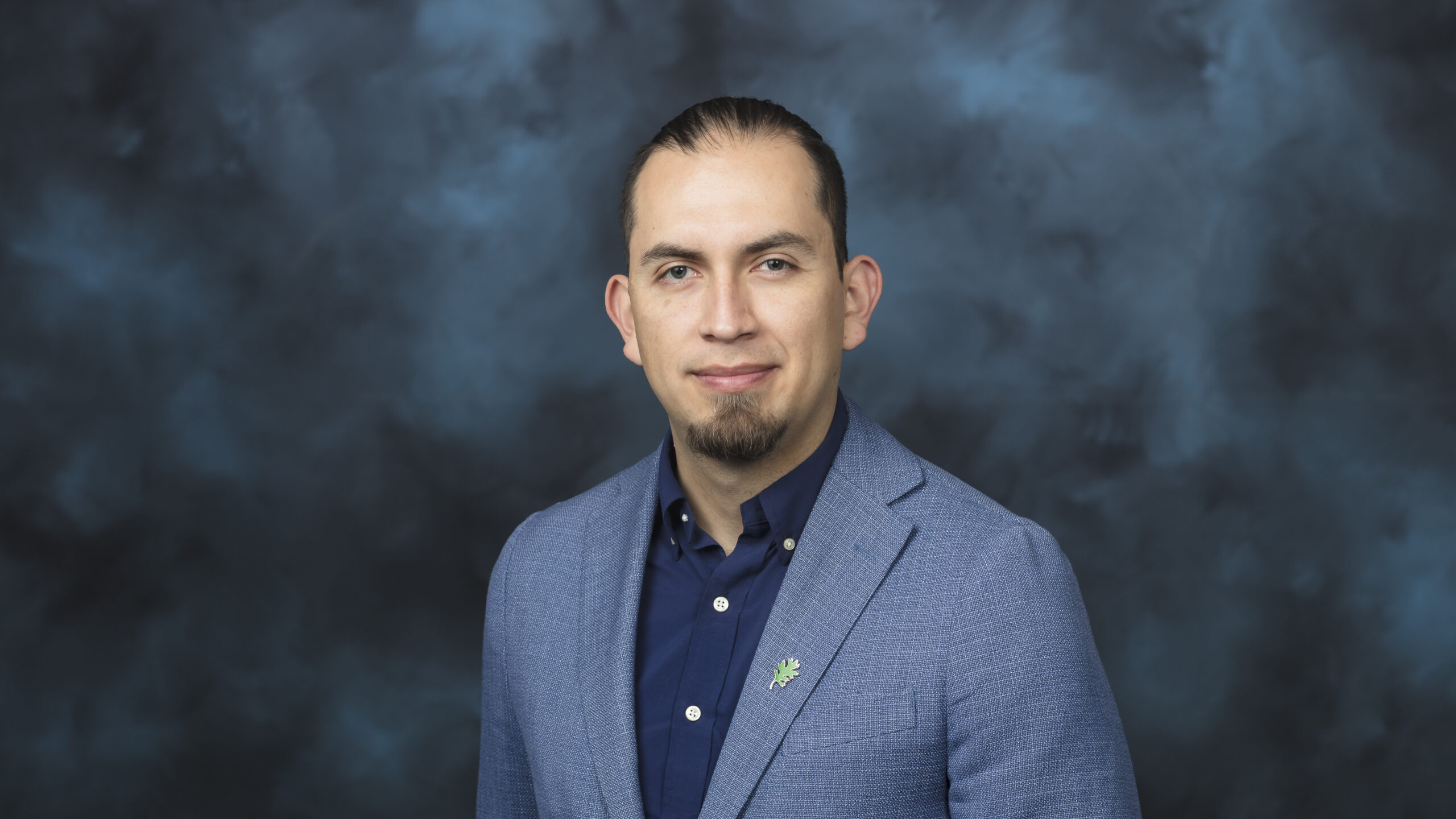 Professional headshot of a man with short dark hair and a goatee wearing a light blue blazer and dark shirt with a small green pin, against a blue-gray background.