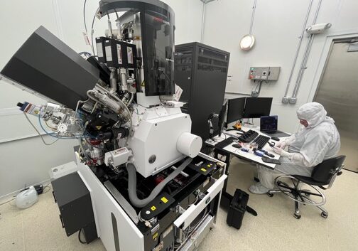 A scientist in a full-coverage white lab suit sits next to a large black and white instrument with protrusions