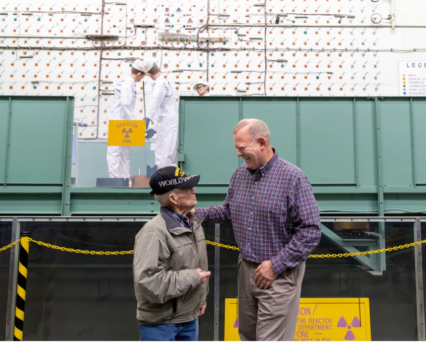 An older man and another man smile and talk in front of a reactor exhibit at ORNL.