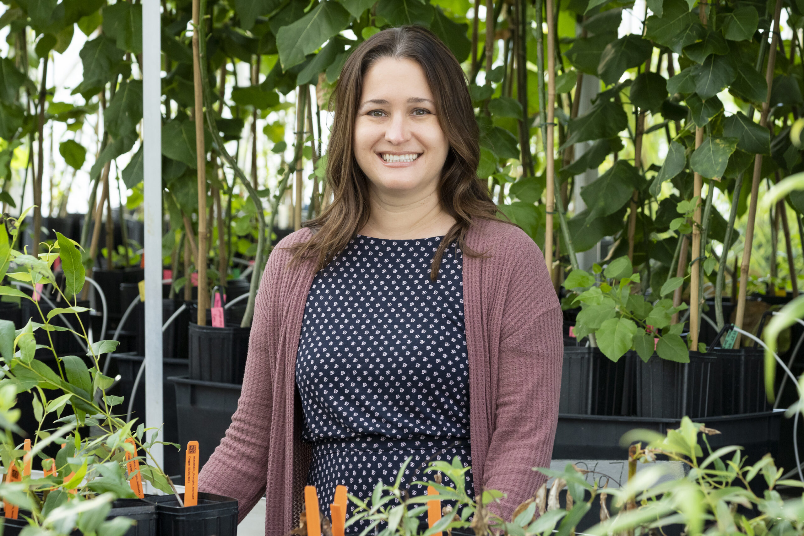 Smiling woman standing in a greenhouse surrounded by potted plants and tall leafy trees.