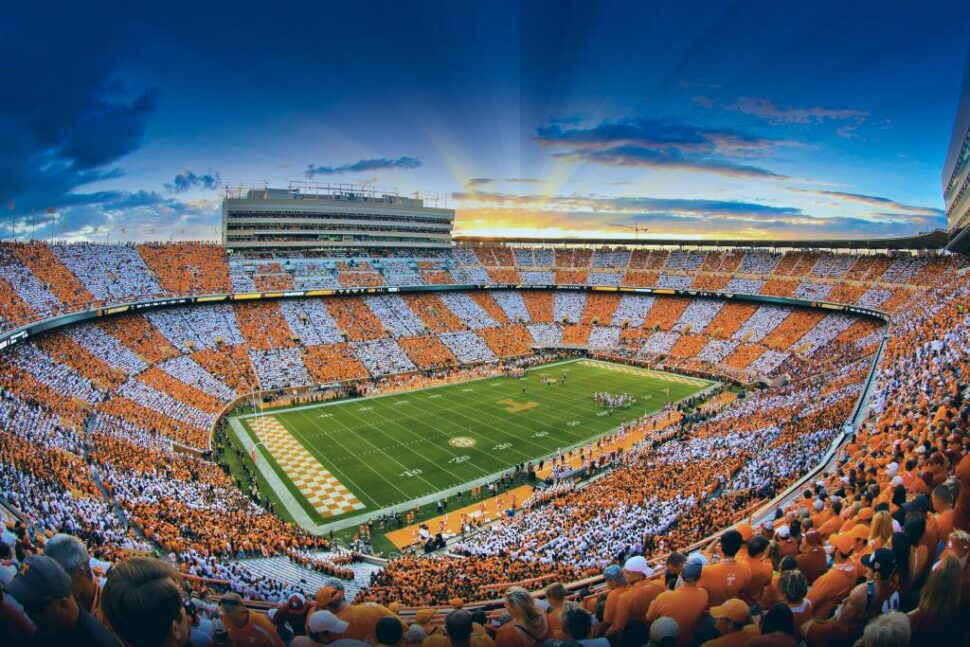 A panoramic view of a packed Neyland football stadium in Knoxville, Tennessee at sunset, with fans dressed in orange and white forming a checkerboard pattern across the stands.