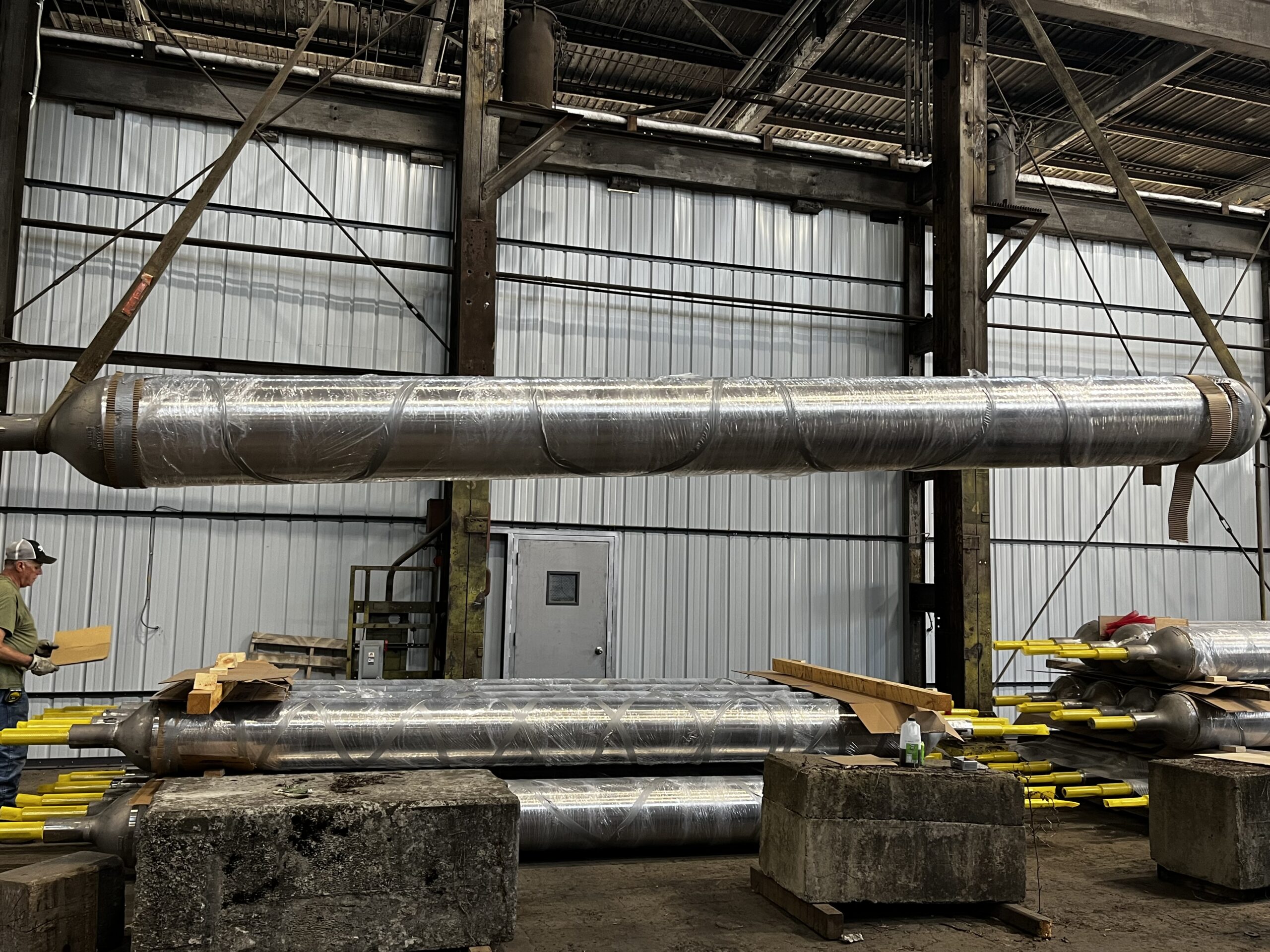 Long cylindrical metal components wrapped in protective plastic are suspended and stacked inside an industrial workshop, with steel beams, concrete blocks, and a worker nearby for scale.