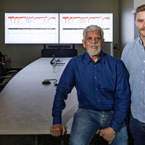 Luiz Leal and Jesse Brown stand in a conference room at Oak Ridge National Laboratory with research data displayed on two monitors behind them. Leal sits on the edge of a conference table while Brown stands beside him during a discussion about nuclear data research.