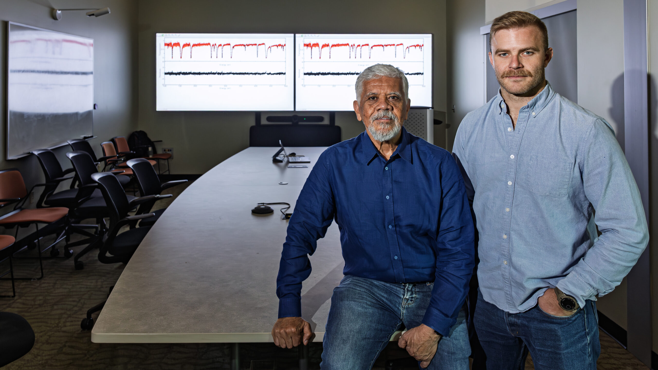 Two researchers pose in a conference room with scientific data displayed on monitors behind them.