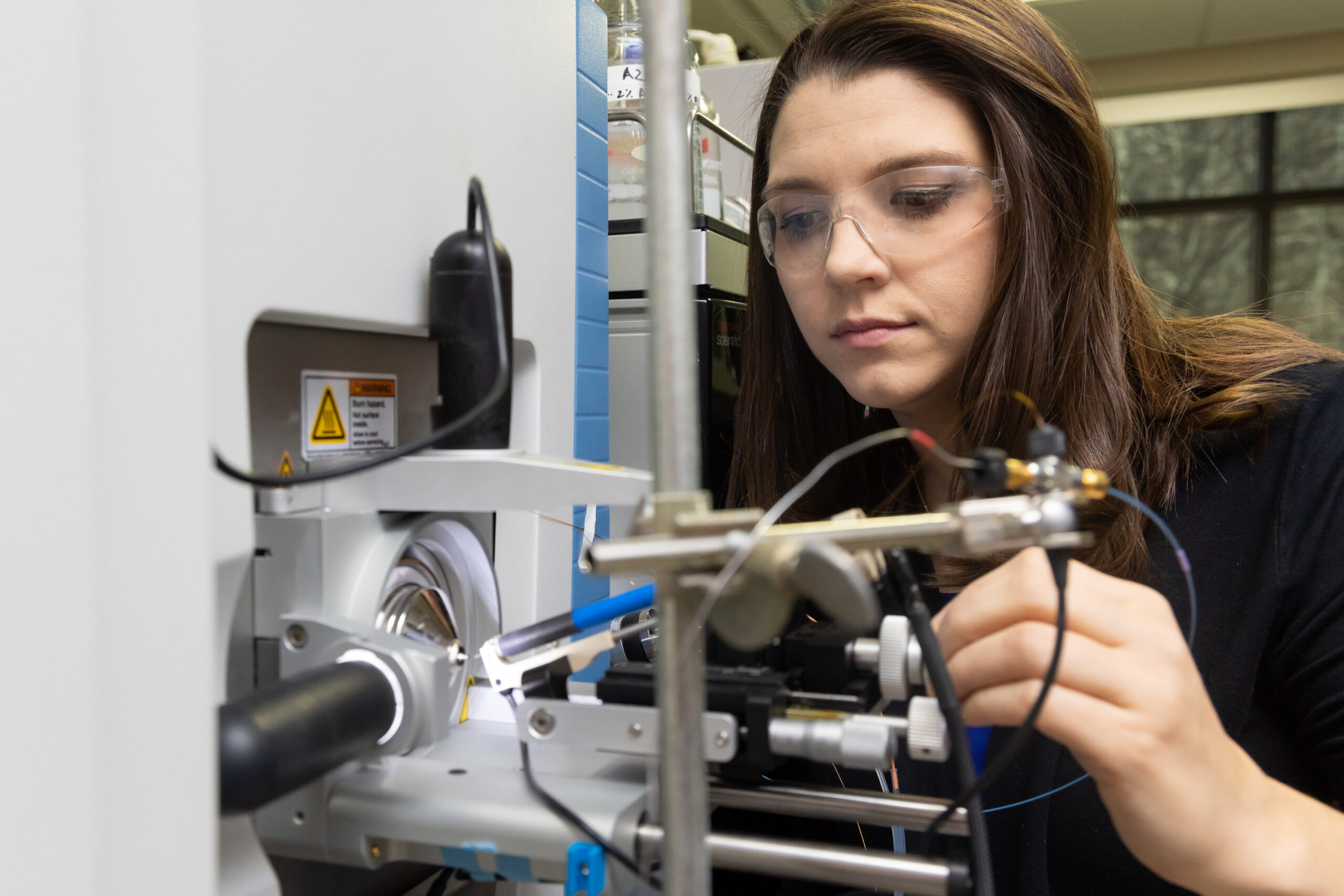 A researcher wearing safety glasses adjusts a precision instrument in a laboratory, focusing on a small component mounted on a metal apparatus surrounded by scientific equipment.