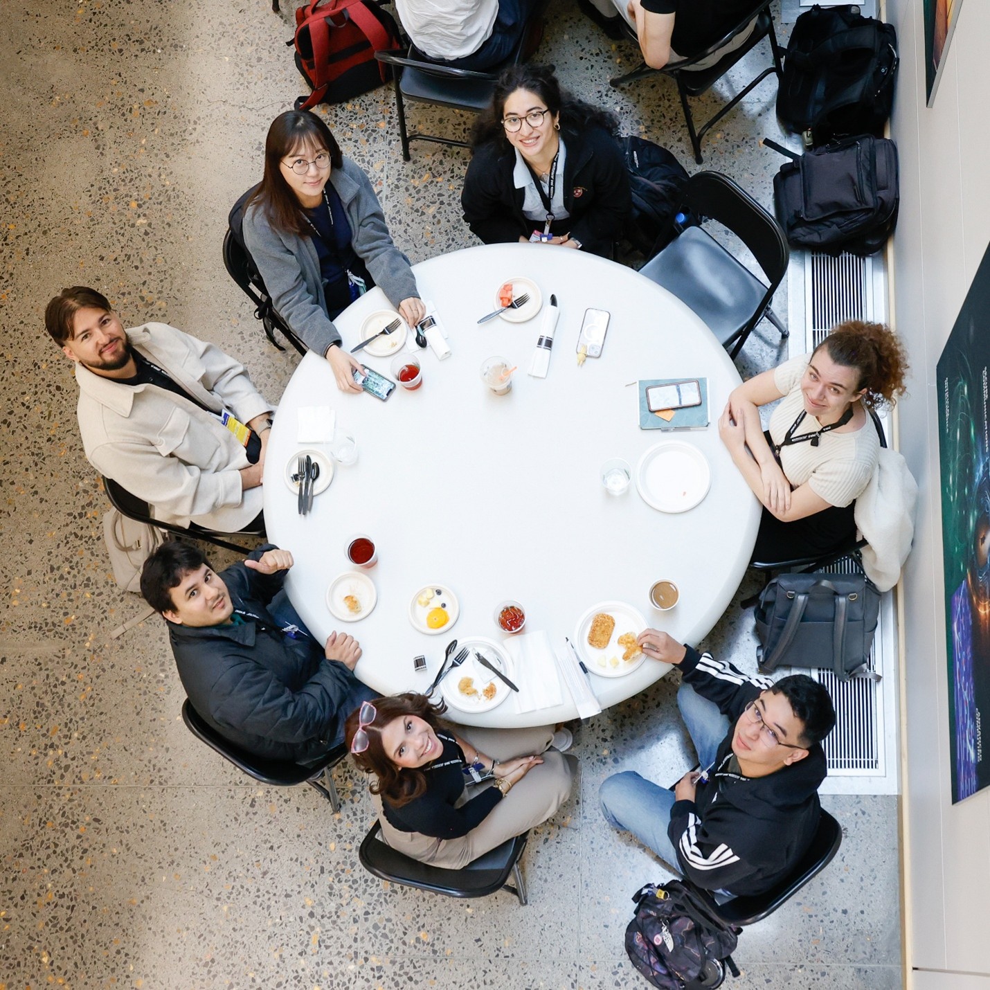 A group of students sit around a round white table, smiling up at the camera during a lunch break, with plates of food and drinks in front of them.