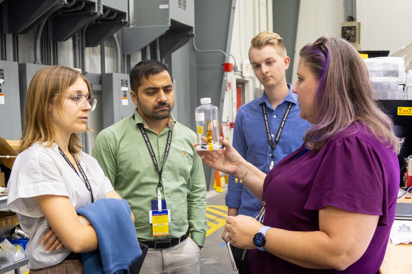 Four students listen attentively as a scientist holds up a clear plastic bottle containing a labeled sample during a tour inside a laboratory facility.