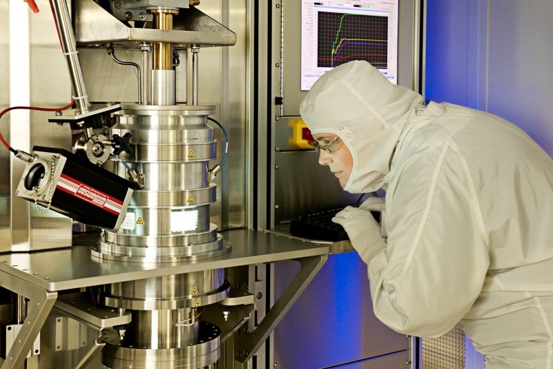 A scientist in a full-coverage white protective suit leans toward a large metal instrument, peering inside