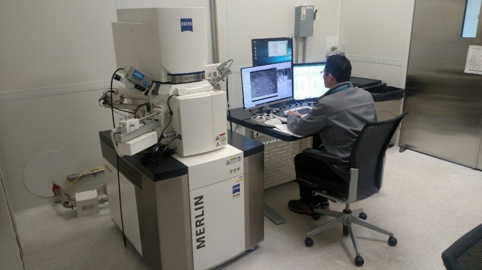 Aman sits at a computer next to a refridgerator-sized machine with small protrusions