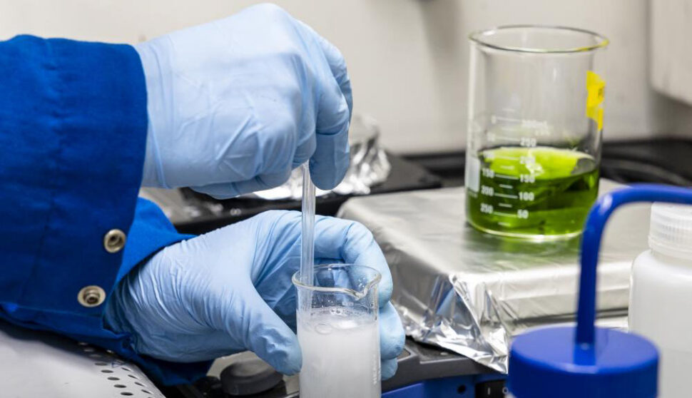 A set of gloved hands manipulates a liquid in a beaker within a lab setting.