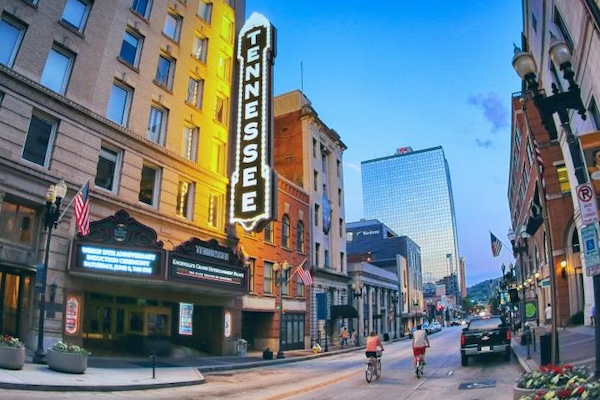 Downtown Knoxville's Gay Street at dusk, featuring the illuminated vertical Tennessee Theatre marquee on the left. Cyclists and vehicles travel along the street, which is lined with historic buildings, storefronts, flags, and streetlights. Modern office towers rise in the background beneath a blue evening sky.