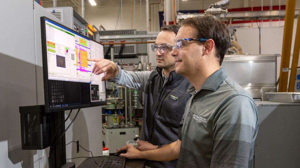 Two scientists wearing safety goggles study a screen together in a lab setting.