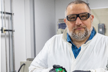 A researcher wearing a lab coat and safety goggles smiles.