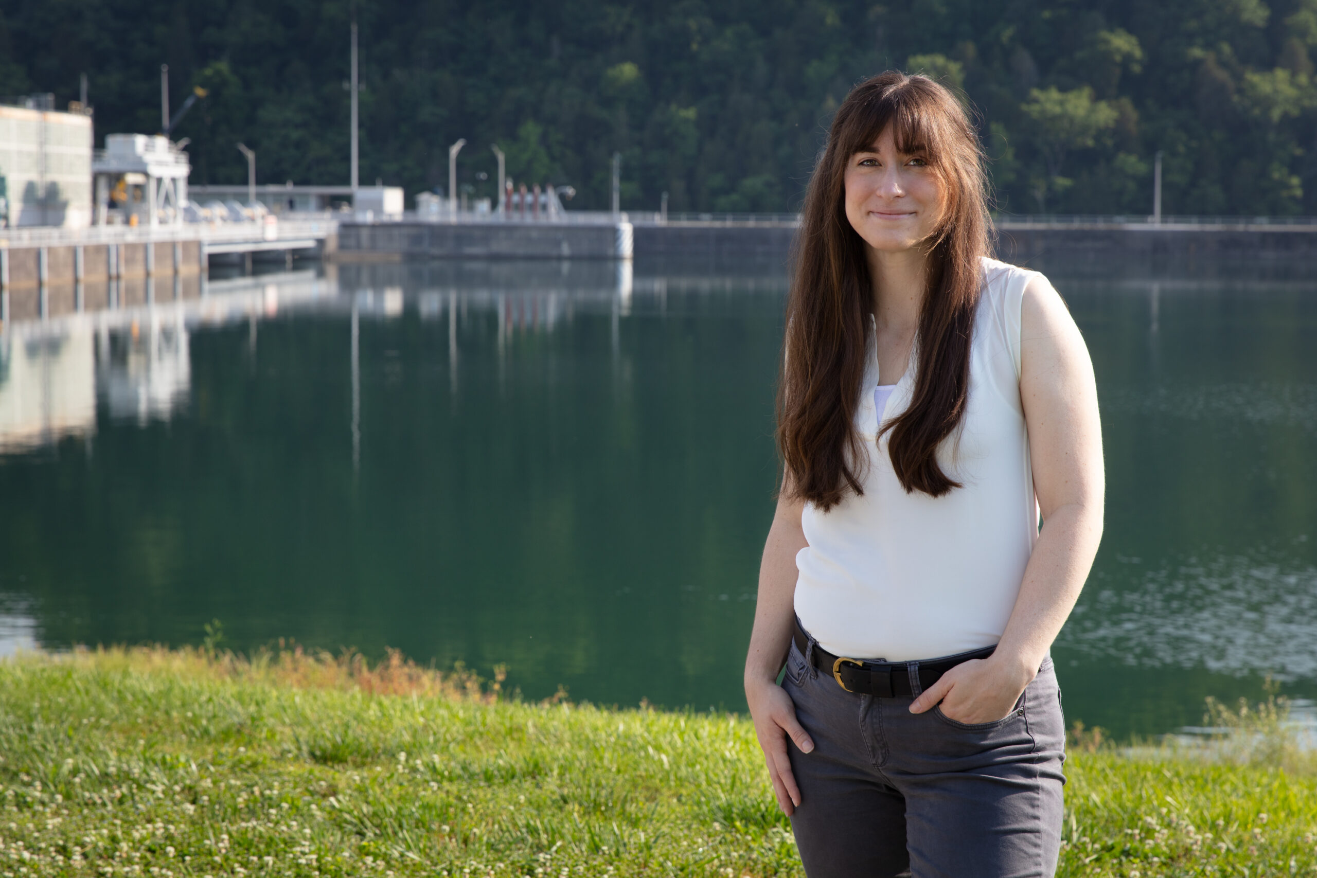 Person standing on a grassy shoreline with a calm reservoir and industrial facility in the background.
