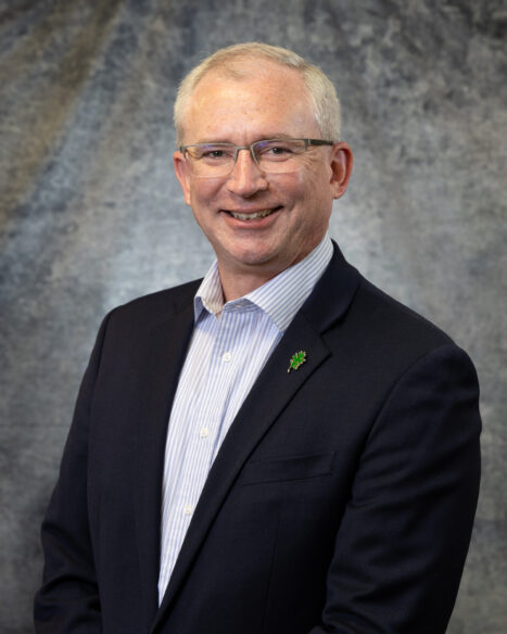 Studio portrait of Brian Kaul, R&D Staff Member at Oak Ridge National Laboratory, wearing a dark suit and light blue shirt while standing in front of a gray textured backdrop at the National Transportation Research Center.