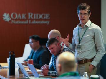 ORNL researcher speaks to colleagues during a meeting while others sit at a conference table using laptops.