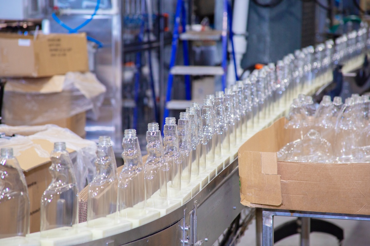 Rows of clear bottles stand neatly on a conveyor belt. They are being transported along a smooth, gray automated system in a bright industrial facility. Cardboard boxes are stacked nearby, and machinery can be seen in the background of the factory. Image from Envato