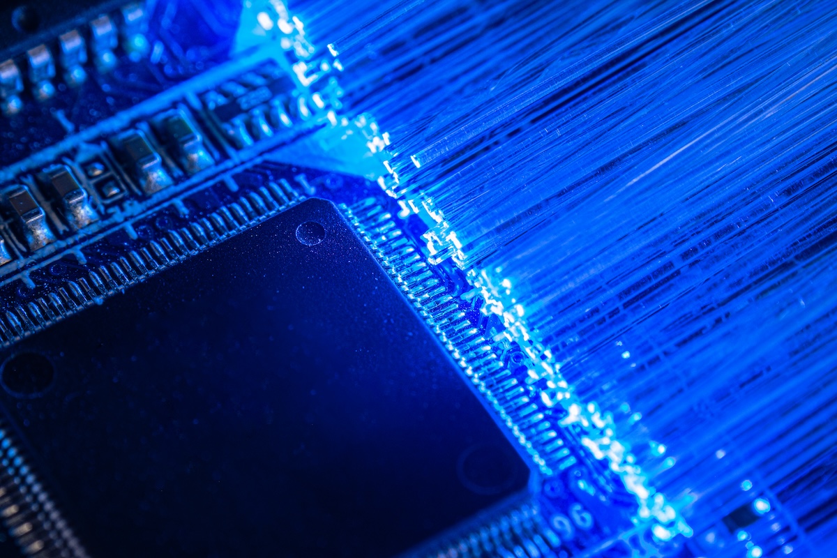 This macro shot shows illuminated fiber optic cables plugged into a black circuit board. The blue light from the cables catches the edges of the board and the circuitry is visible on the sides of the board. Image from Envato