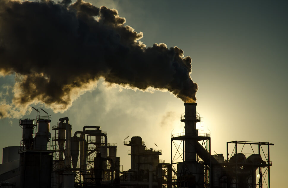 Silhouetted industrial complex against a hazy sky at sunset. The image features a cluster of industrial structures with various towers, pipes, and buildings. Thick, dark smoke billows out of a tall chimney, obscuring the sun and creating a dramatic contrast against the sky. Image from Envato.