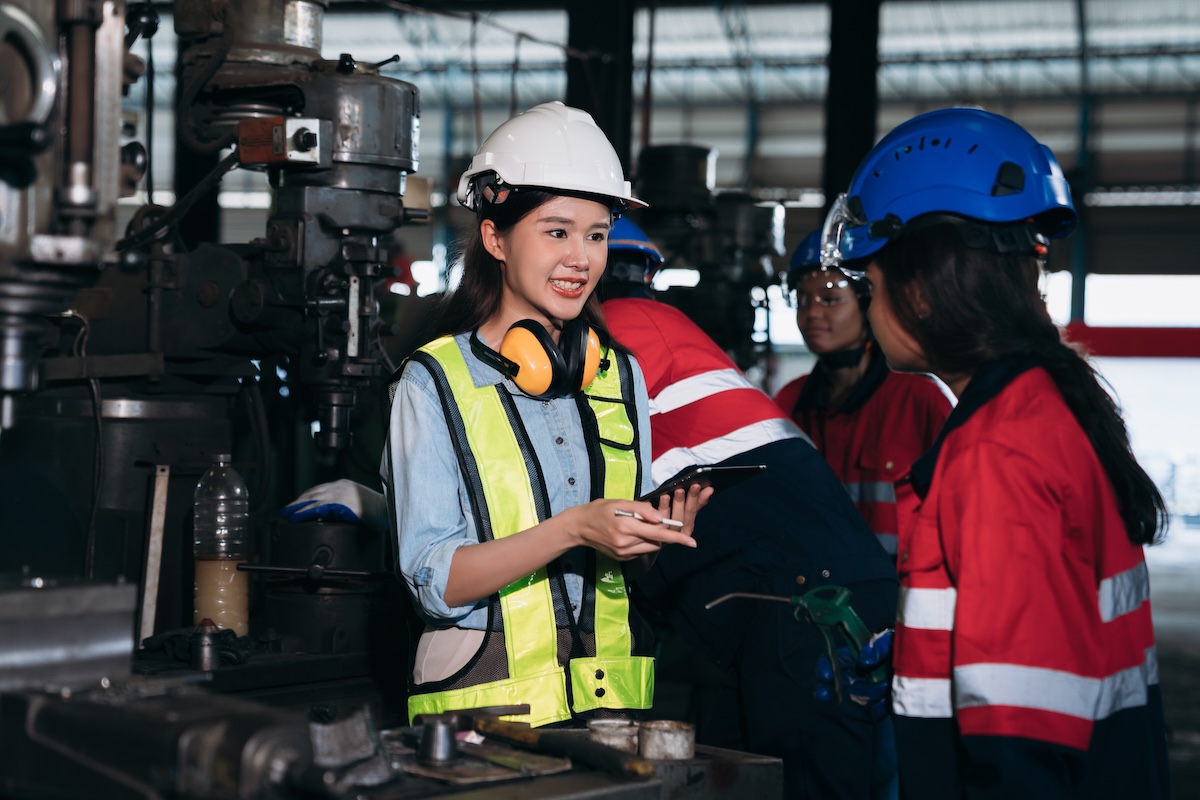 Three women and one other person wearing helmets and safety gear are gathered near machinery in a factory setting. One woman is holding a tablet and gesturing with a pen, while another is listening attentively. Image from Envato
