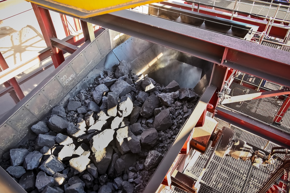 A top-down shot reveals a bin full of jagged, dark grey rocks within an industrial setting. The rocks vary in size, filling most of the frame. The bin is constructed of dark metal with red support beams. Image from Envato