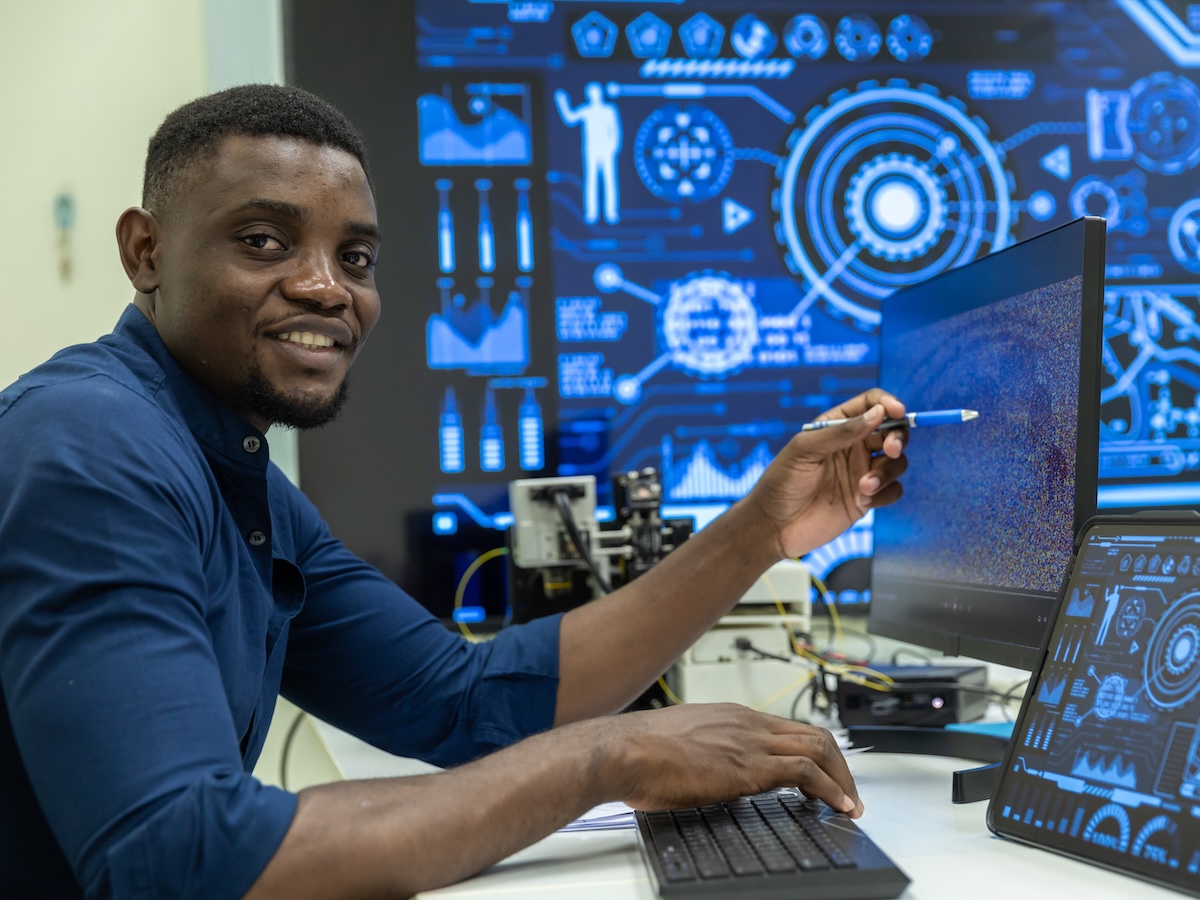 A smiling adult man with short dark textured hair sits at a desk with a computer, keyboard and tablet. He wears a blue button down shirt. The background features a large screen displaying digital, technical images with blueprints and graphics. Image from Envato.