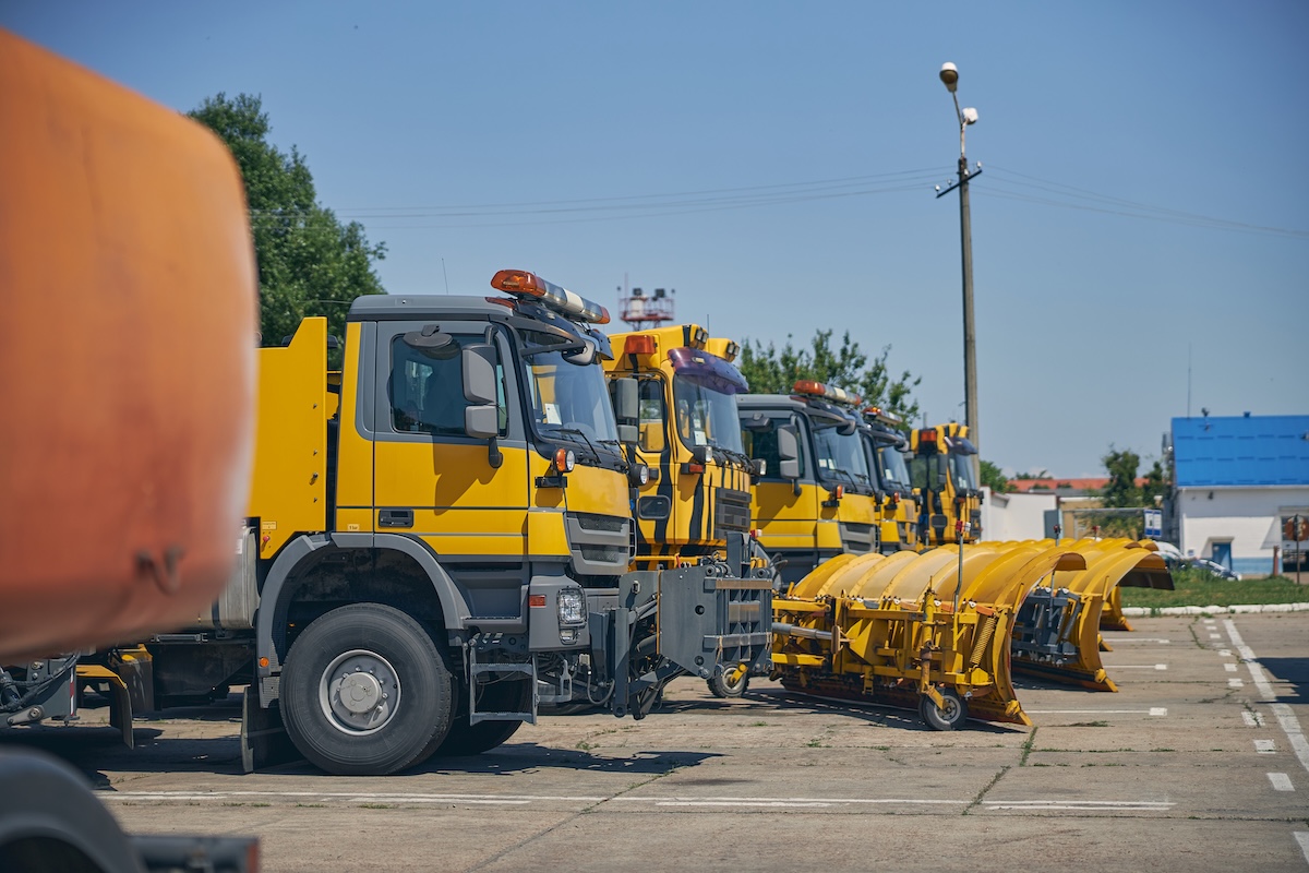 A line of yellow and grey snowplow trucks sit parked on a paved lot under a bright blue sky. The trucks are equipped with snowplow blades. One of the trucks has an orange tank or container. The setting is urban with buildings and a utility pole visible. Image from Envato