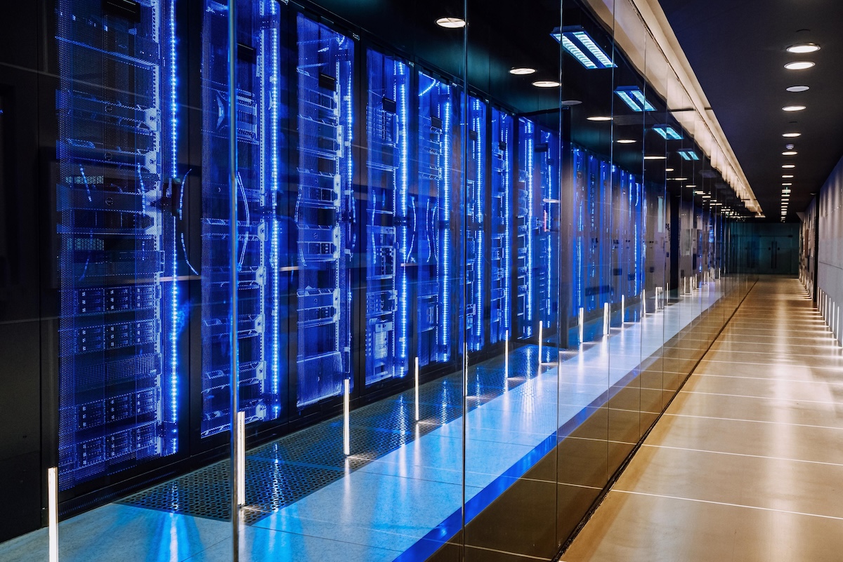 An eye-level view of a server room. Numerous data servers are visible behind glass, illuminated by glowing blue lights. Image from Envato