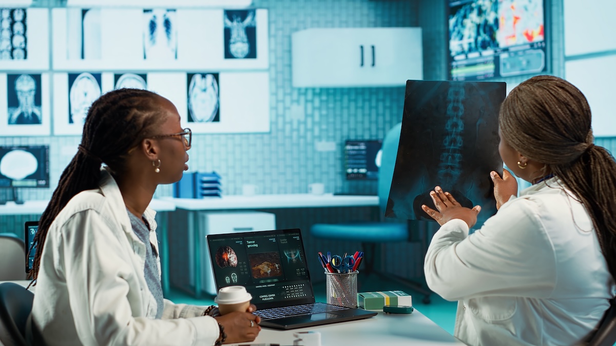 Two women in white coats at a light blue-walled hospital workplace setting analyze medical data. One woman with glasses and dark braided hair holds a coffee cup and sits at a table with a laptop showing medical imagery, while the other doctor stands and gestures toward a spinal X-ray film. Image from Envato
