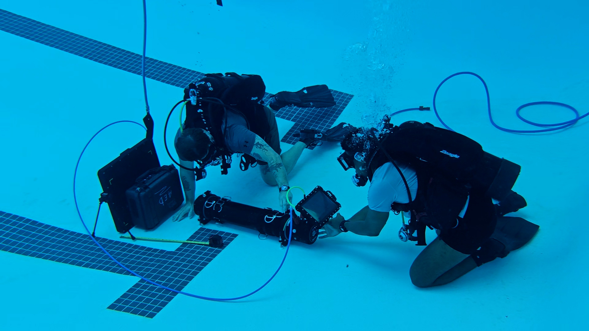 Two divers in scuba gear work underwater in a pool, examining and adjusting equipment connected by cables on the pool floor.