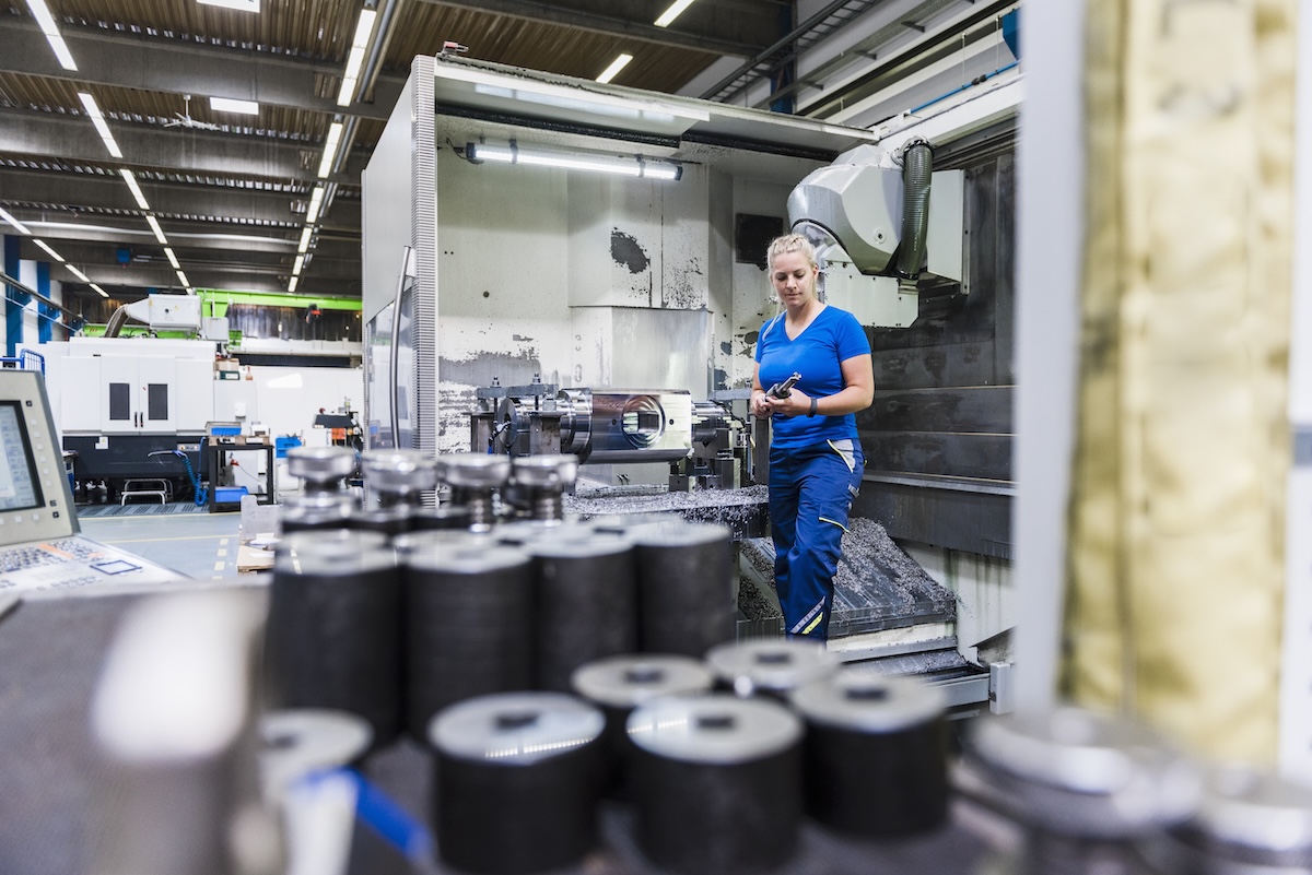 A woman with her hair in a bun wearing a blue shirt and matching trousers, stands inside a metal milling machine in an industrial setting. The setting is a factory floor. In the foreground are rows of metal tools. Image from Enavto