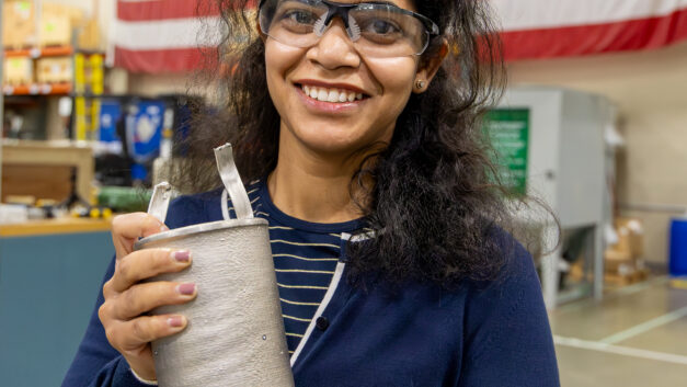 ORNL researcher Vanshika Singh holds an electroformed nickel component developed for advanced nuclear energy manufacturing research while standing in front of a large American flag.