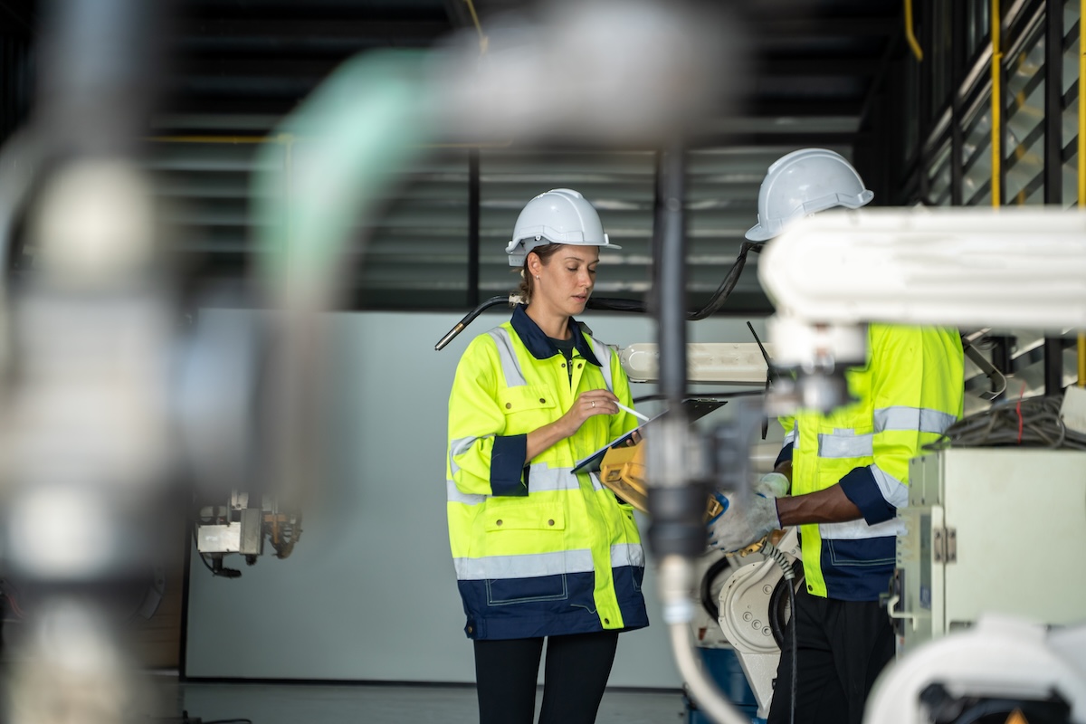 Two people wearing hard hats and high visibility jackets are seen in a manufacturing setting. The fair haired woman has a tablet in hand. Industrial machinery is visible. Image from Envato