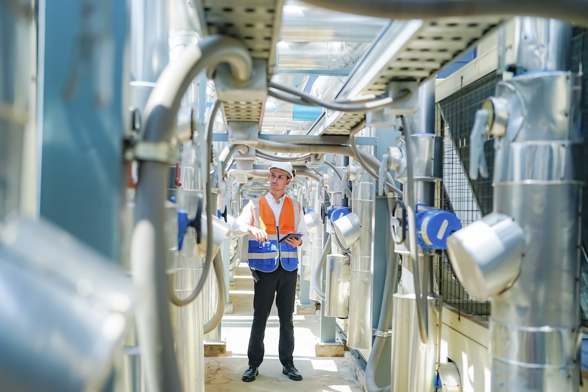 A serious adult man with short dark hair is seen wearing a white hard hat, white button-down shirt, black pants, and black shoes. He also wears an orange and blue safety vest, while holding a dark tablet, as he stands and examines the complex arrangement of industrial pipes, cables, and silver tanks within a workplace setting. Image from Envato