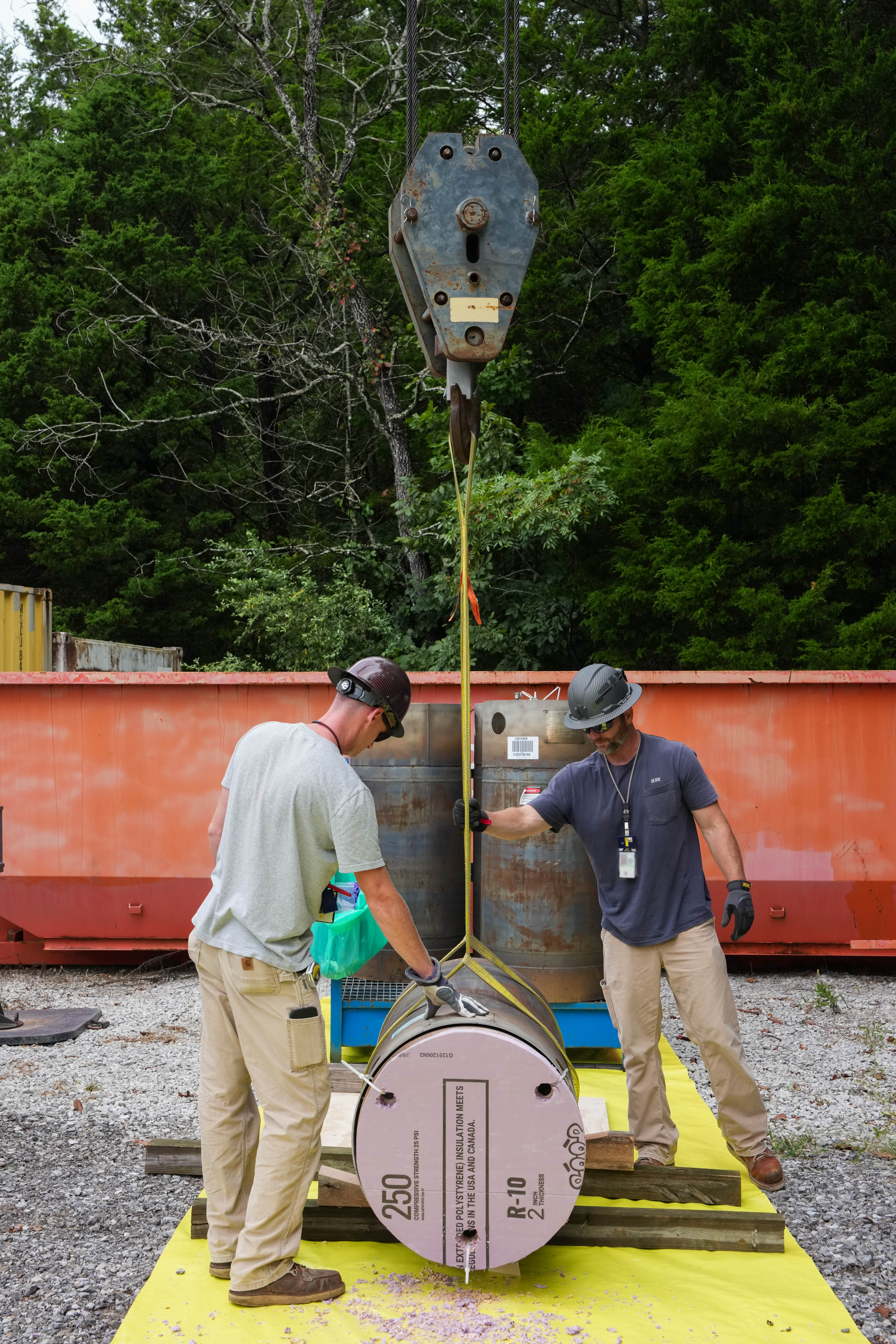 Two workers guide a cylindrical component suspended from a crane.