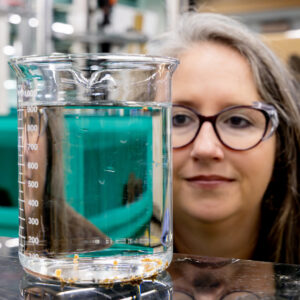 Kristine Moody holds and examines a large laboratory beaker filled with water and polymer samples during materials testing in a laboratory at Oak Ridge National Laboratory.